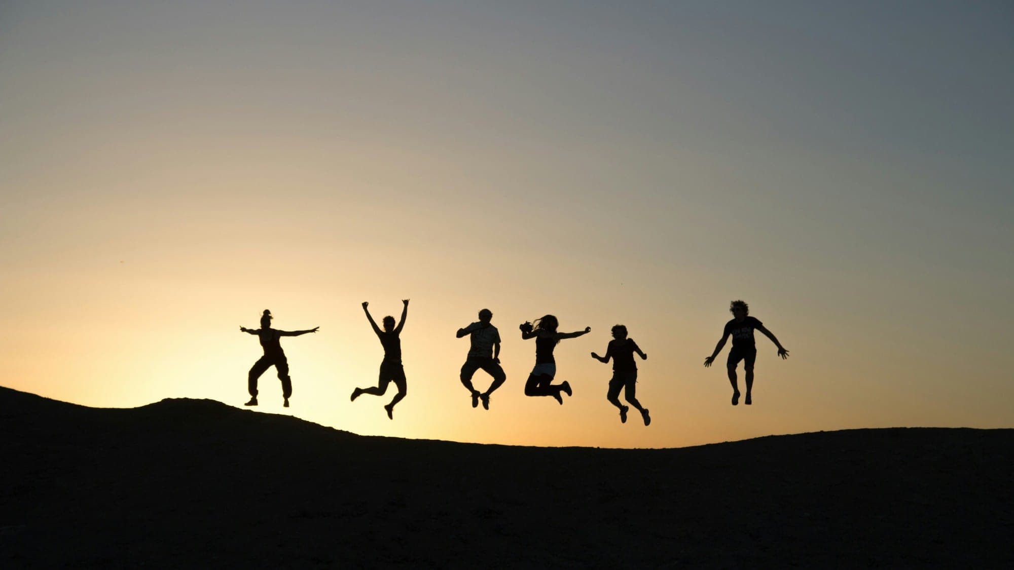 Six people jumping for joy on a hilltop, silhouetted against a golden sunset sky