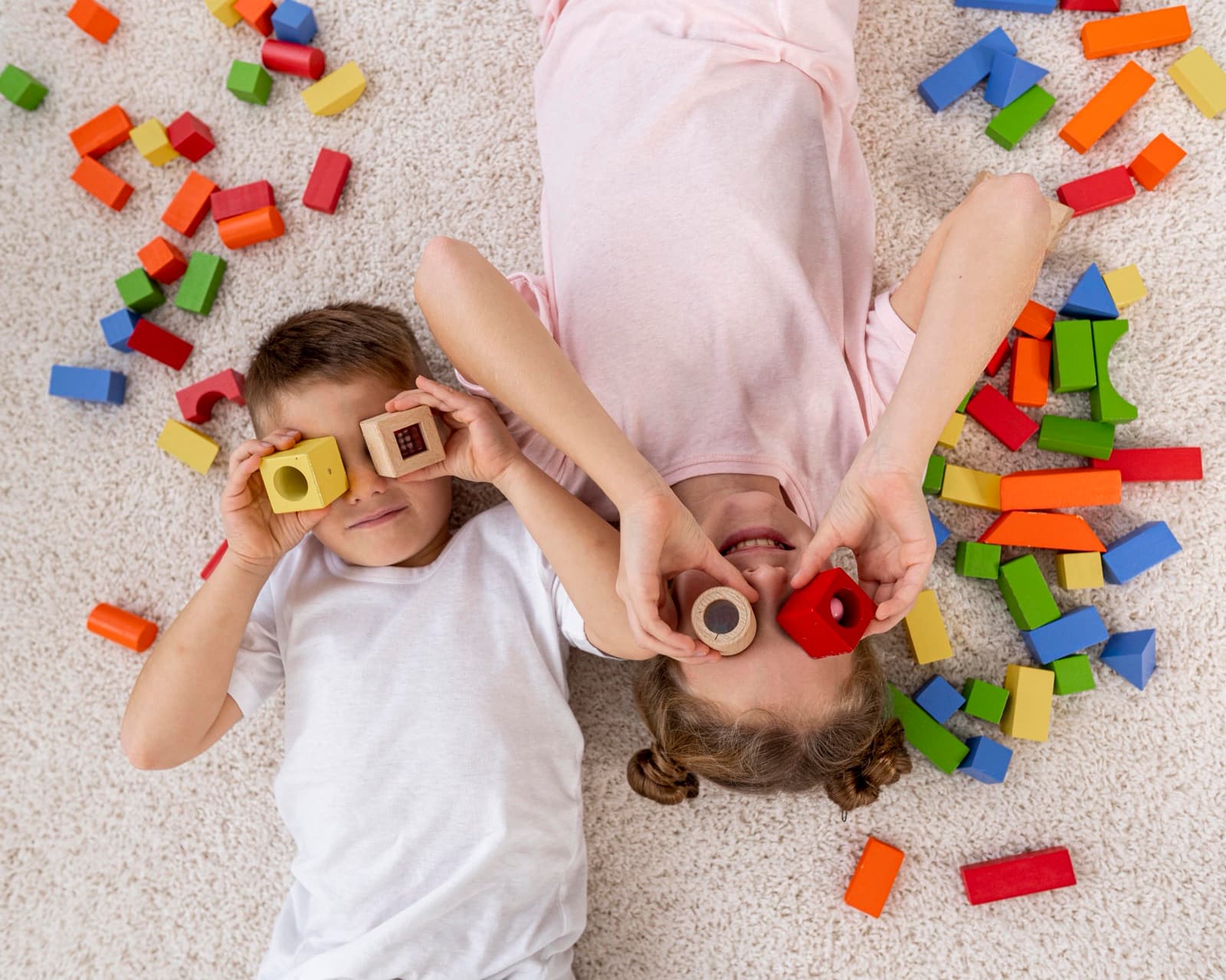 Two children lying on a carpet, playing with colourful wooden blocks